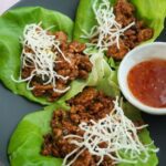 Ground chicken on top of green lettuce with white rice sticks on top of a black plate.