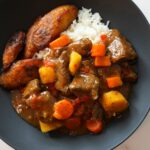 Beef stew for two in a black bowl with white rice and golden plantain on the side.