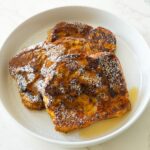 Pumpkin French toast on a white plate, topped with maple syrup, whipped honey butter, and powdered sugar.