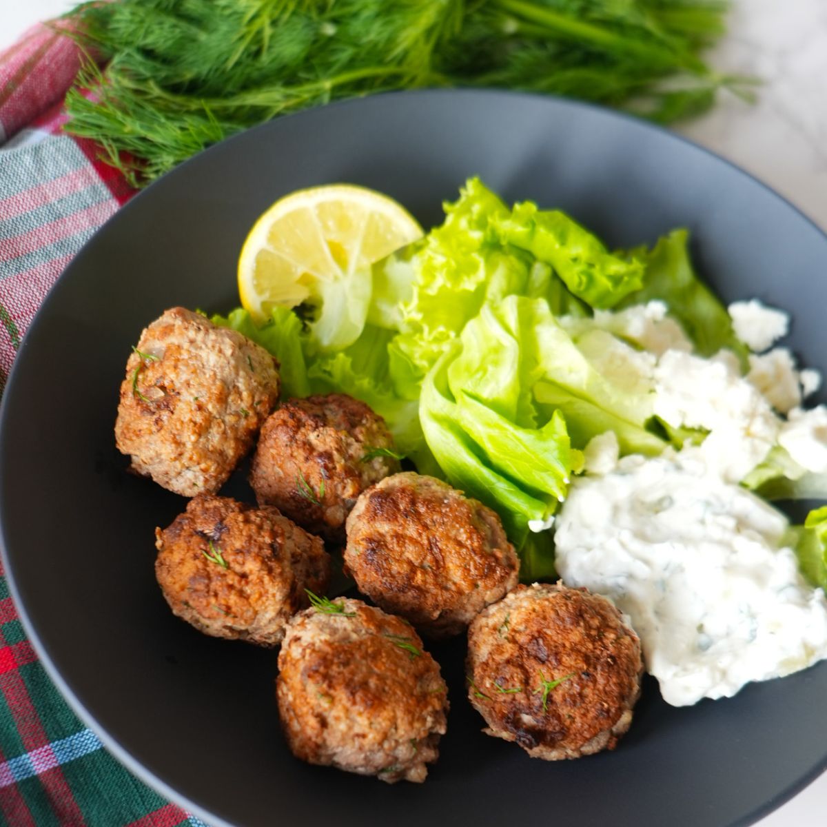 Greek turkey meatballs with tzatziki sauce, fresh dill garnish, and a side salad on a black plate.