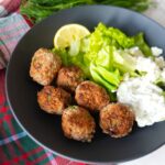 Greek turkey meatballs with tzatziki sauce, fresh dill garnish, and a side salad on a black plate.
