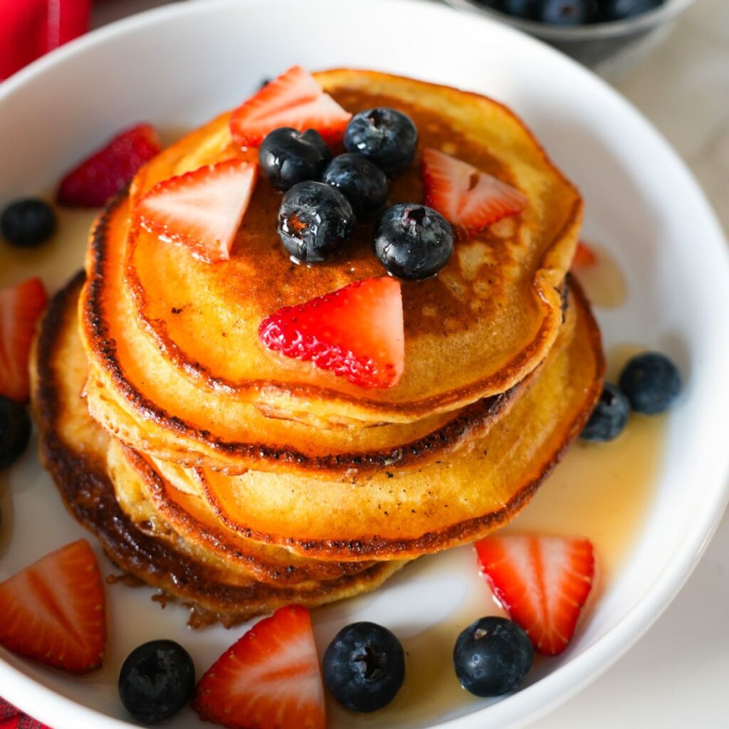 A stack of fluffy lemon ricotta pancakes topped with blueberries, strawberries and drizzled with maple syrup.