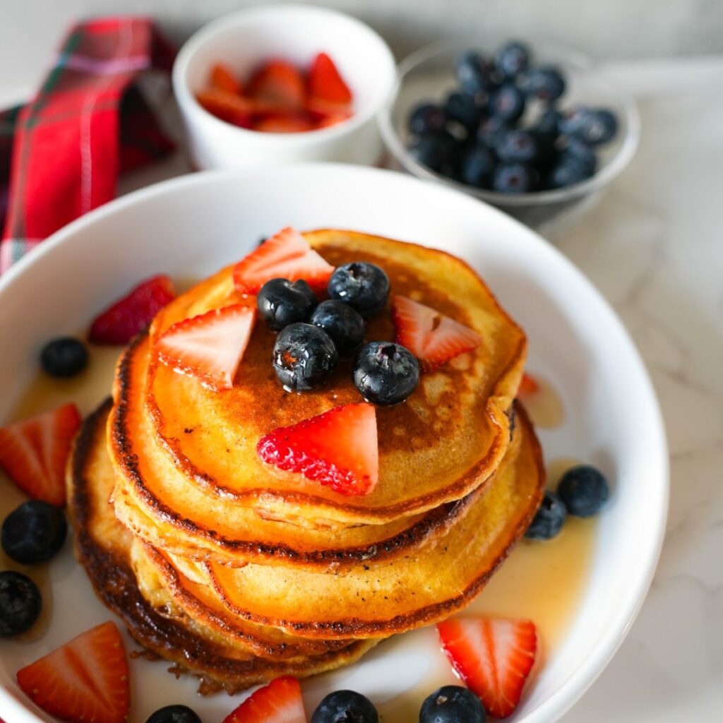 A stack of fluffy lemon ricotta pancakes topped with blueberries, strawberries and drizzled with maple syrup.
