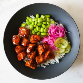 Teriyaki salmon on top of rice with pickled cucumber and onions in a black bowl.