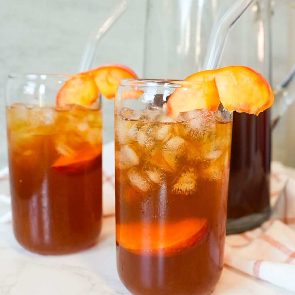 Two glasses of sweet peach tea with ice and fresh peach slices on top.