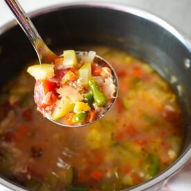 Chicken vegetable soup with shredded chicken, colorful vegetables and broth in a ladle with a pot in the back.
