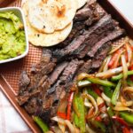 Steak fajitas on a sheet pan tray with tortillas and guacamole.