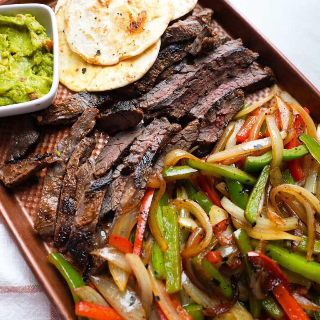 Cooked onions and peppers, skirt steak, guacamole and tortilla shells on a sheet pan.