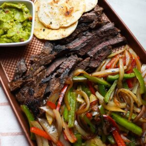 Cooked onions and peppers, skirt steak, guacamole and tortilla shells on a sheet pan.