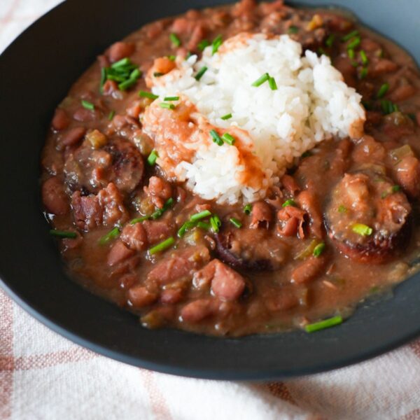Bowl of red beans and rice with sliced sausage, garnished with chopped chives and parsley on top of fluffy white rice.