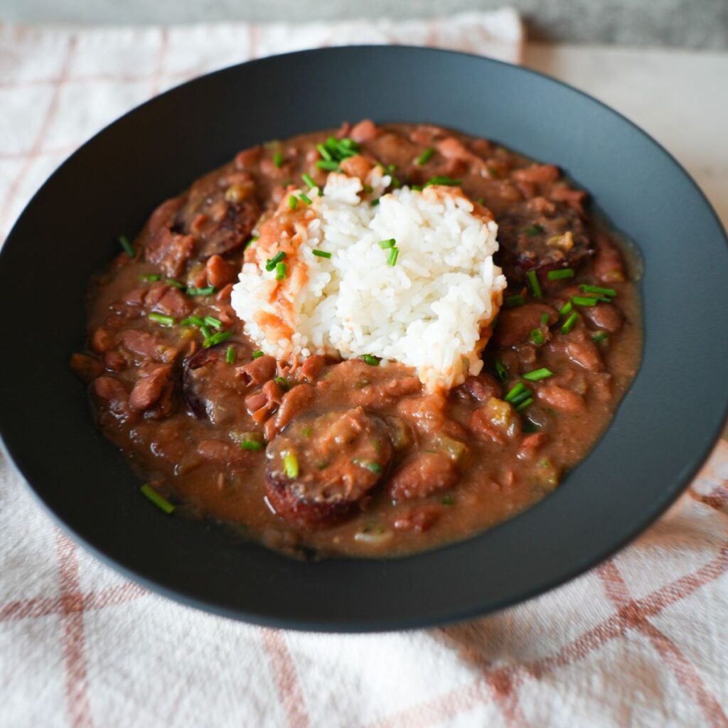 Bowl of red beans and rice with sliced sausage, garnished with chopped chives and parsley on top of fluffy white rice.