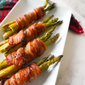Bacon wrapped asparagus bundles baked on a white plate with caramelized edges and brown sugar on top.