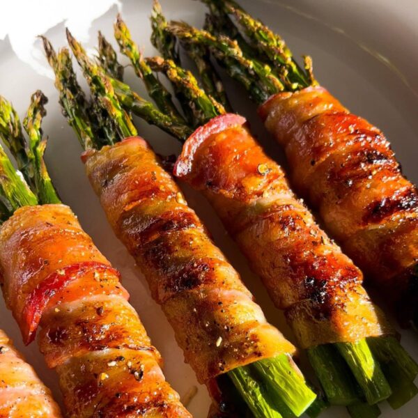 Bacon wrapped asparagus bundles baked on a white plate with caramelized edges and brown sugar on top.