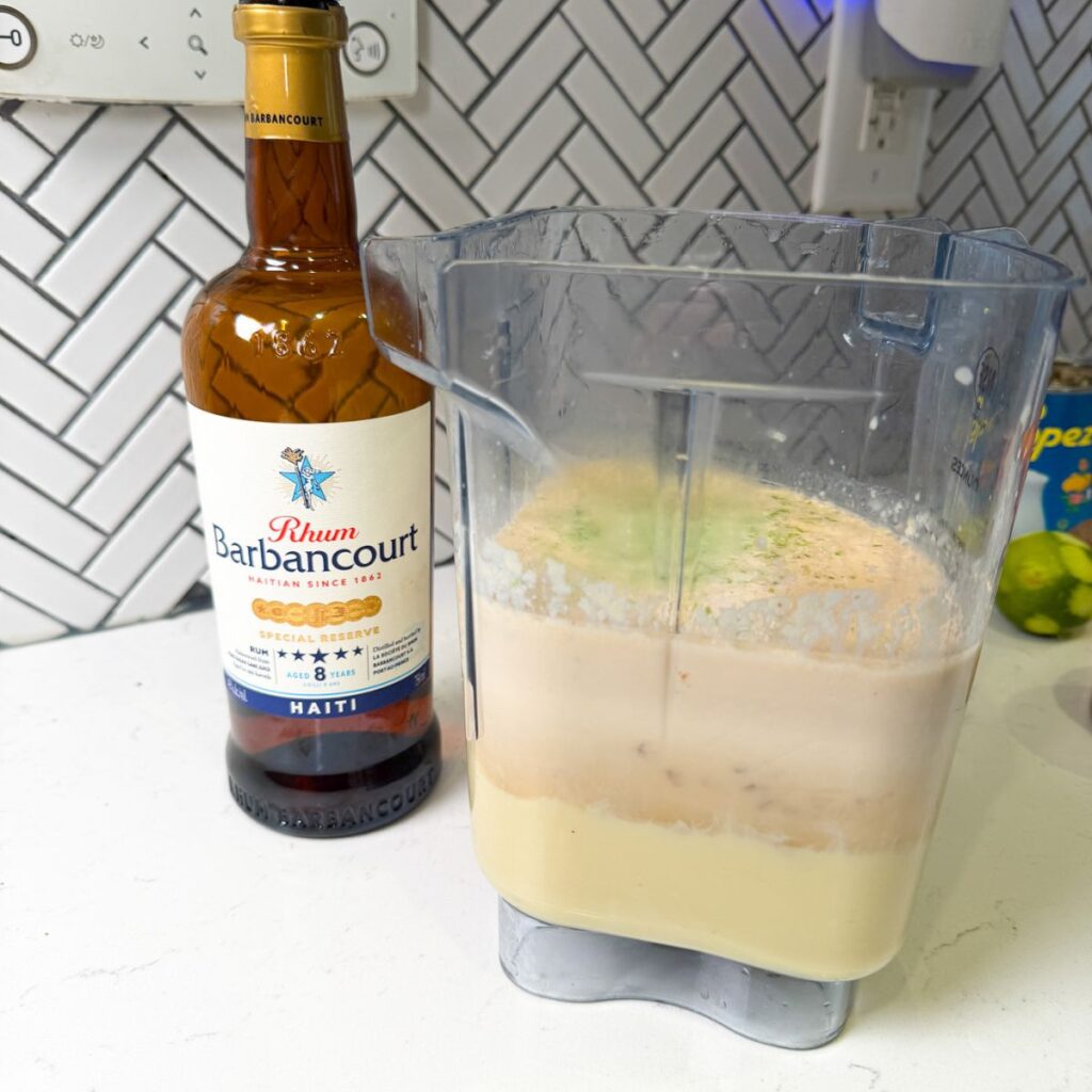 Cremas before blending in a blender next to a bottle of barbancourt rum.