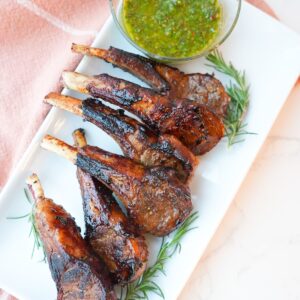Lamb chops with a side of mint chimichurri on a plate with visible herbs and a glossy seared crust.