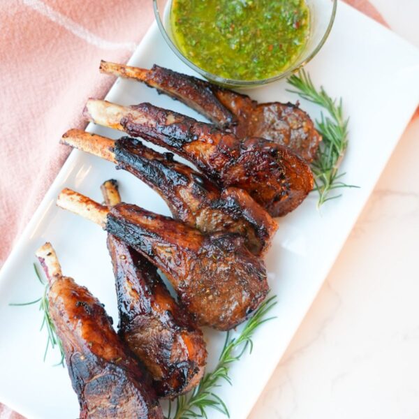 Lamb chops with a side of mint chimichurri on a plate with visible herbs and a glossy seared crust.