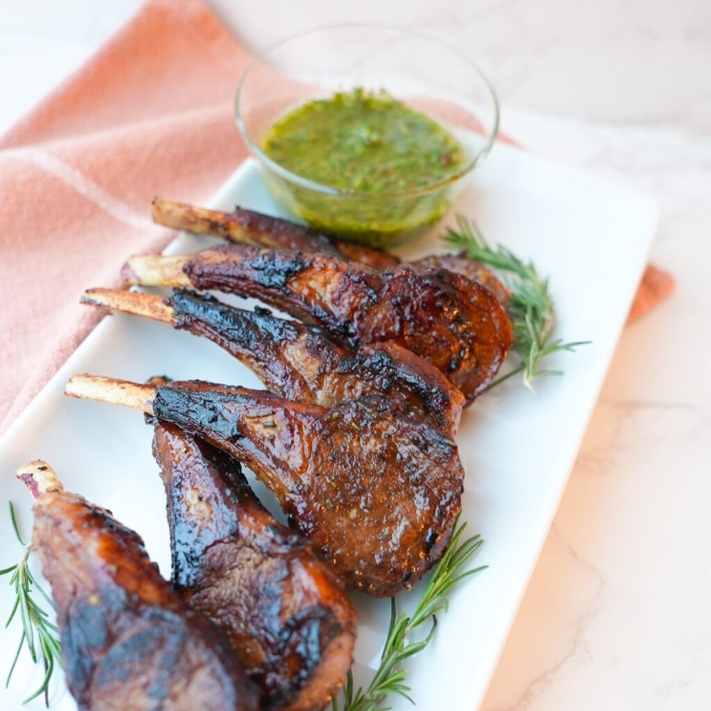 Lamb chops with a side of mint chimichurri on a plate with visible herbs and a glossy seared crust.