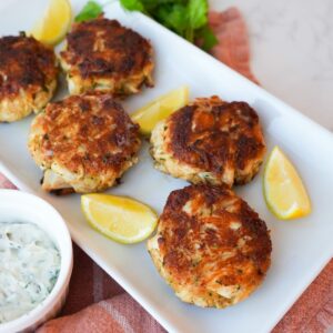 Golden crab cakes on a plate with crispy edges, fresh parsley, and a side of dipping sauce.