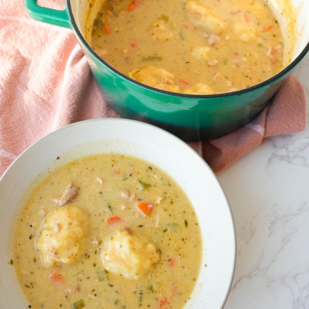 Chicken and dumplings in a bowl and pot with tender chicken, carrots, celery, fluffy dumplings, and creamy broth.