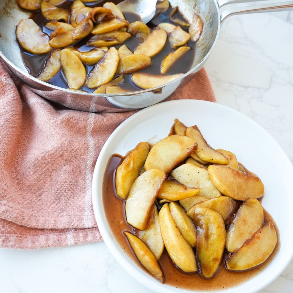 Southern fried apples in a skillet and on a plate, coated in a glossy cinnamon sugar butter sauce.