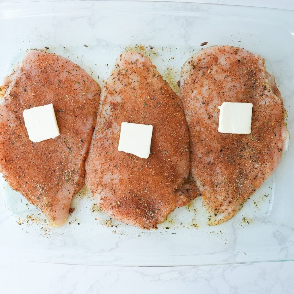 Seasoned raw chicken breast with slices of butter on top.