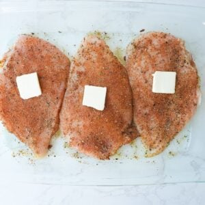Seasoned raw chicken breast with slices of butter on top.